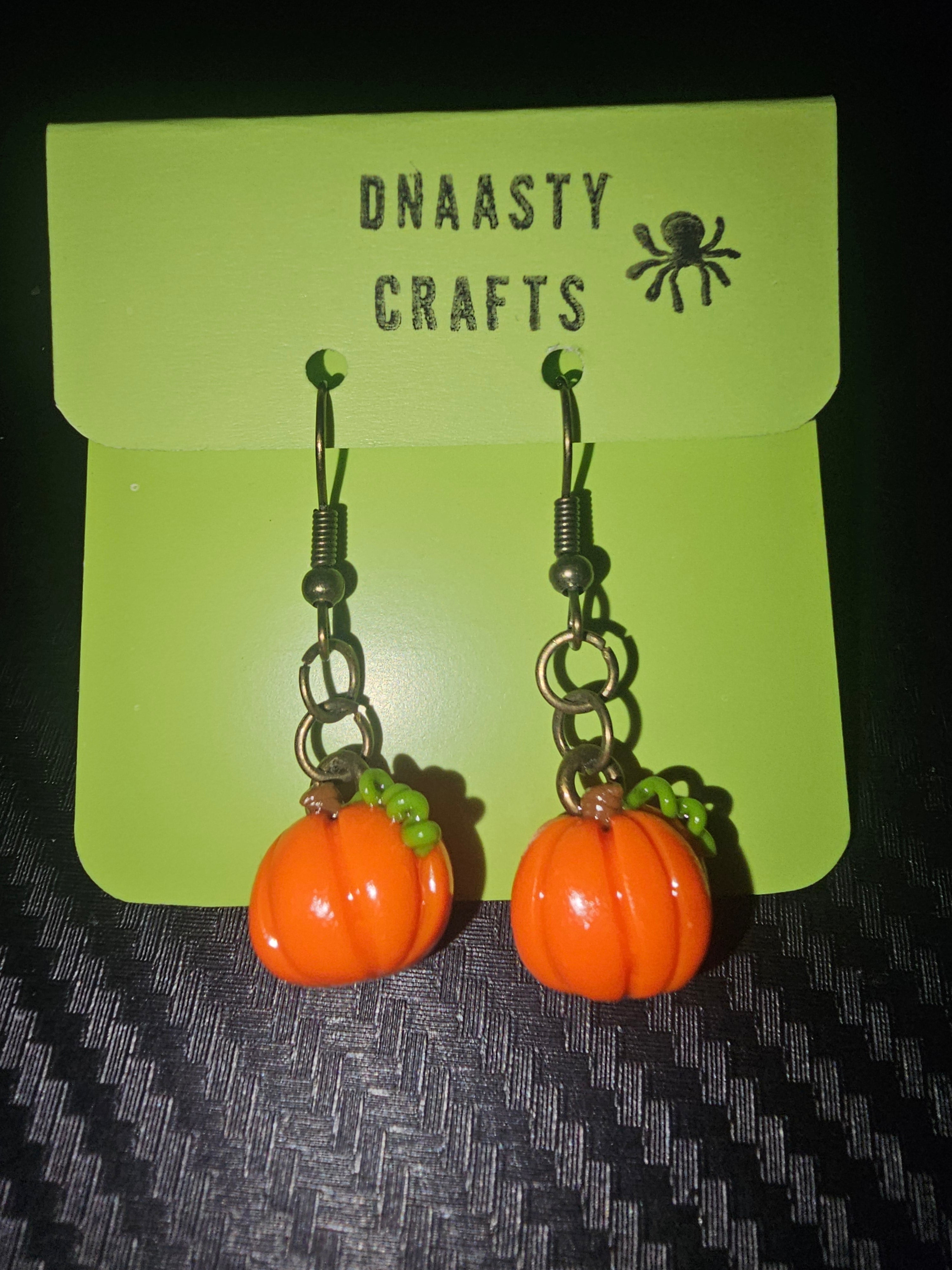 Halloween pumpkin earrings with orange pumpkin charms, green bead accents, and metal hooks, displayed on a Dnaasty Crafts card