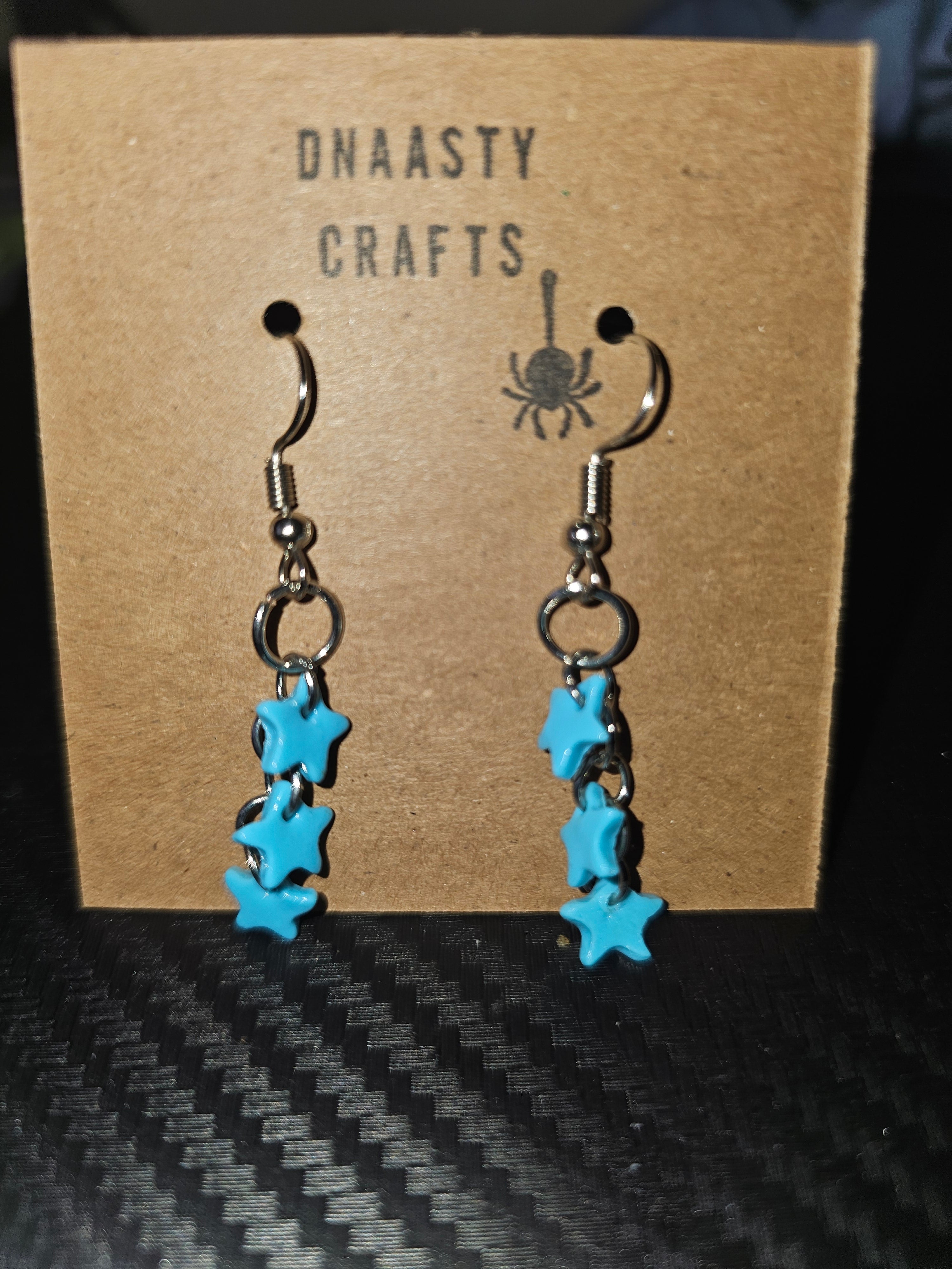 Blue star earrings with polymer clay stars hanging from silver hooks, displayed on a brown Dnaasty Crafts card with black text and spider logo
