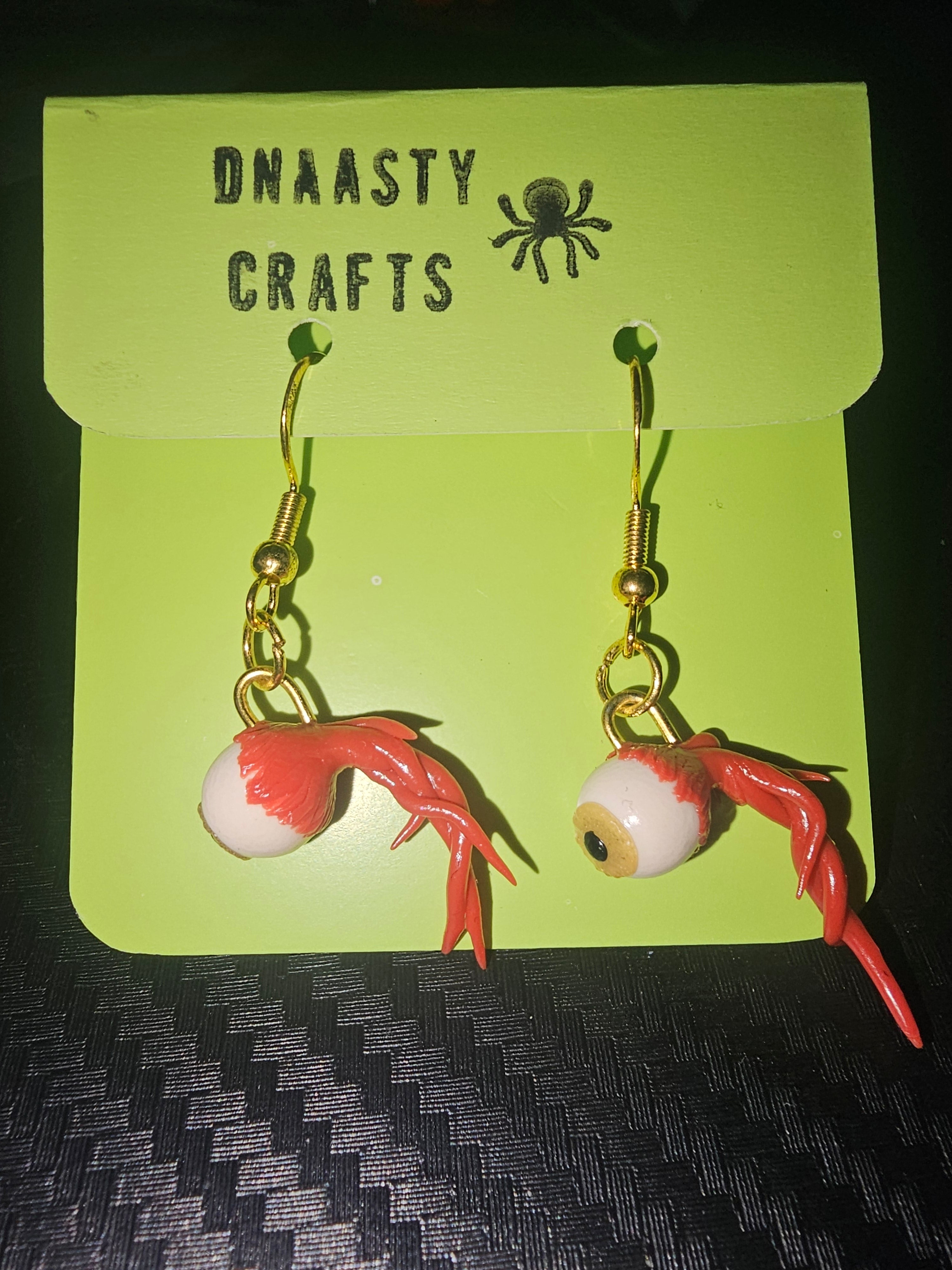 Handmade eyeball earrings featuring realistic white eyeballs with detailed irises, red veins, and gold hooks, crafted from polymer clay, displayed on green branded card with spider logo, perfect for spooky or Halloween-themed jewelry.