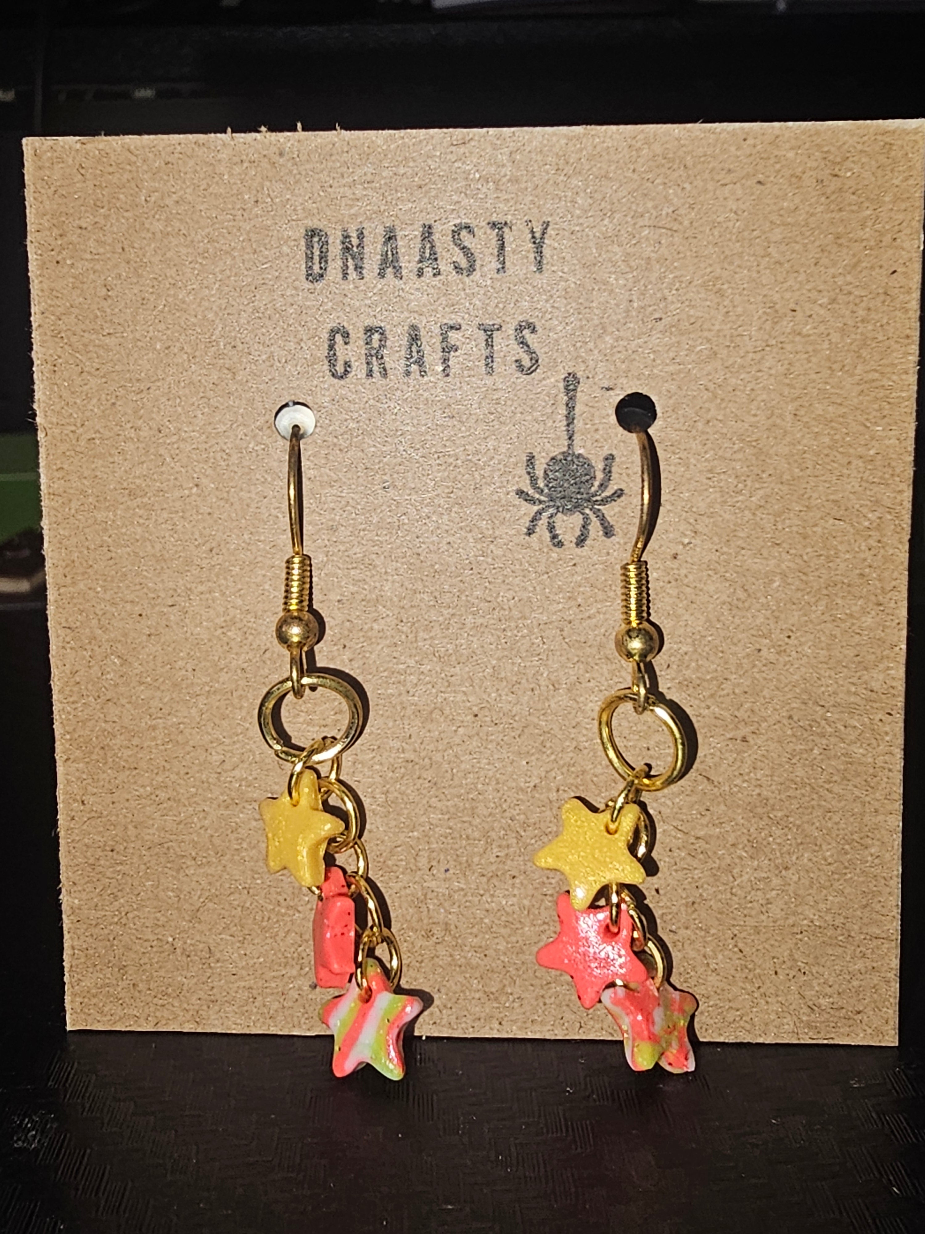 Colorful star-shaped polymer clay earrings with gold hooks hanging on a brown craft display card with 'ONNASTY CRAFTS' and a spider illustration