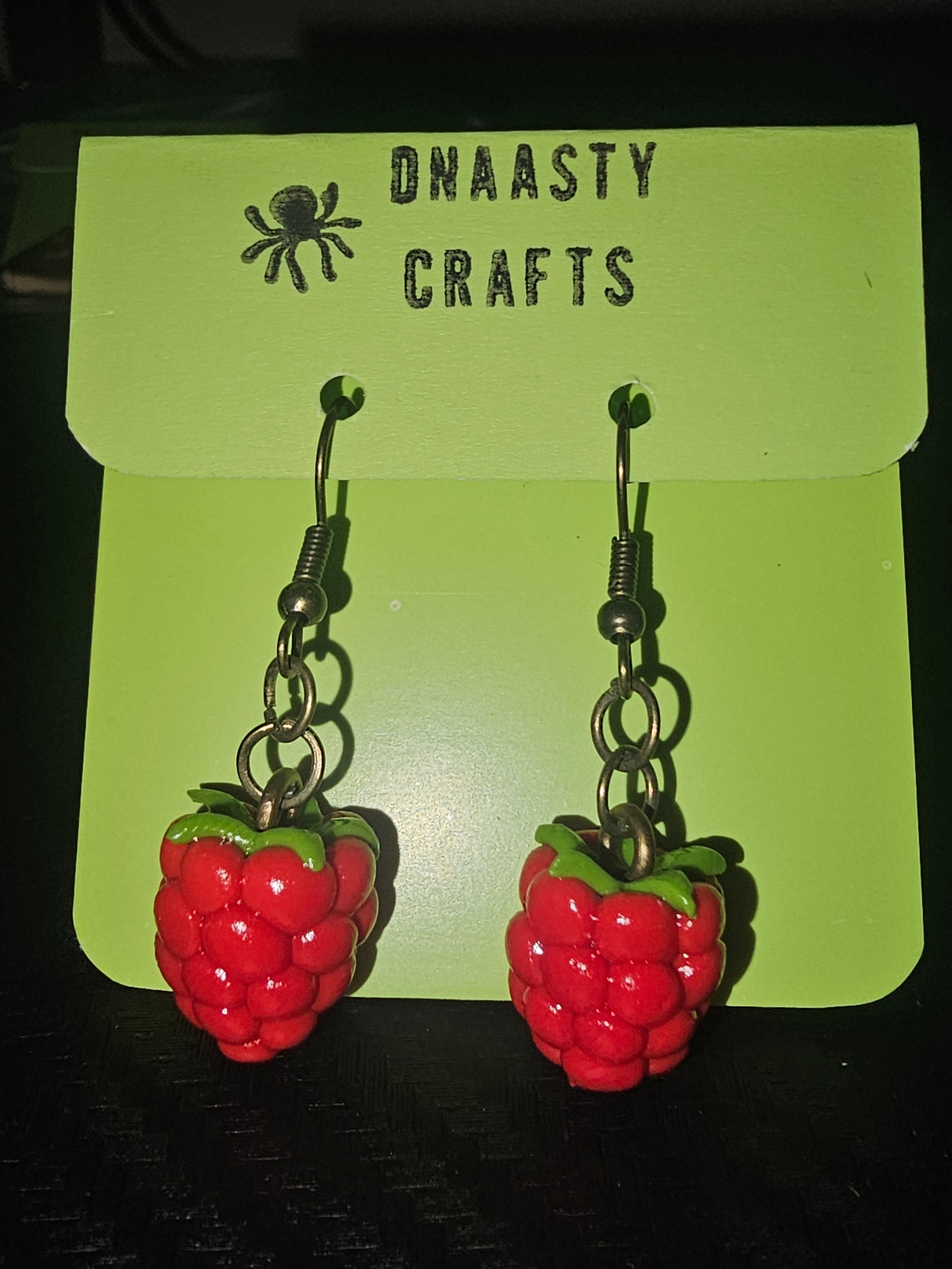 Red and green strawberry-shaped earrings made of glossy polymer clay with metal hooks, displayed on a green Dnaasty Crafts card