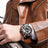 Luxury men's chronograph sports watch with large black dial, rose gold accents, and black silicone strap on a man's wrist, brown leather jacket in background