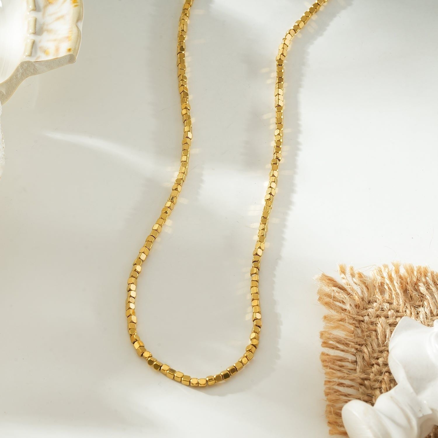Gold beaded dainty chain necklace with small square beads, resting on a decorative ceramic dish with a scalloped edge, ideal for layering or everyday wear