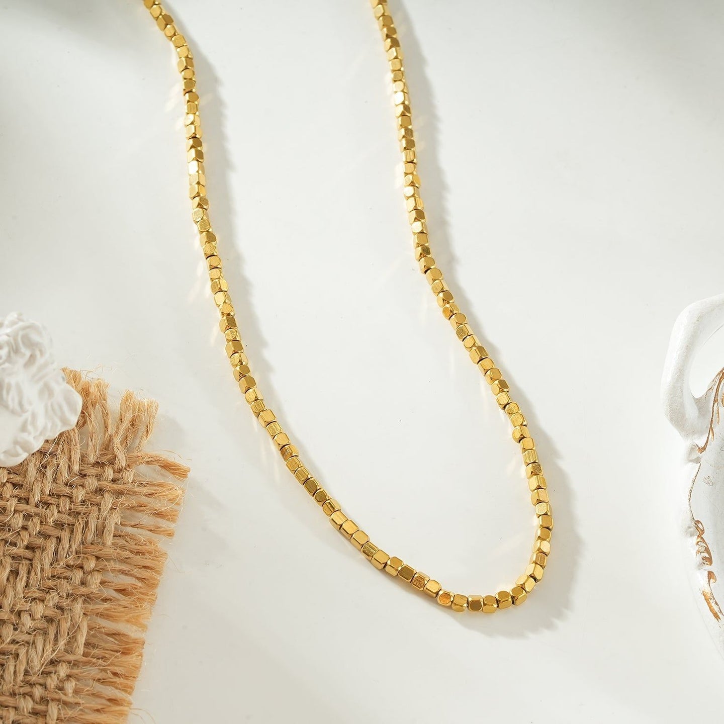 Gold beaded dainty chain necklace with small square beads, resting on a decorative ceramic dish with a scalloped edge, ideal for layering or everyday wear