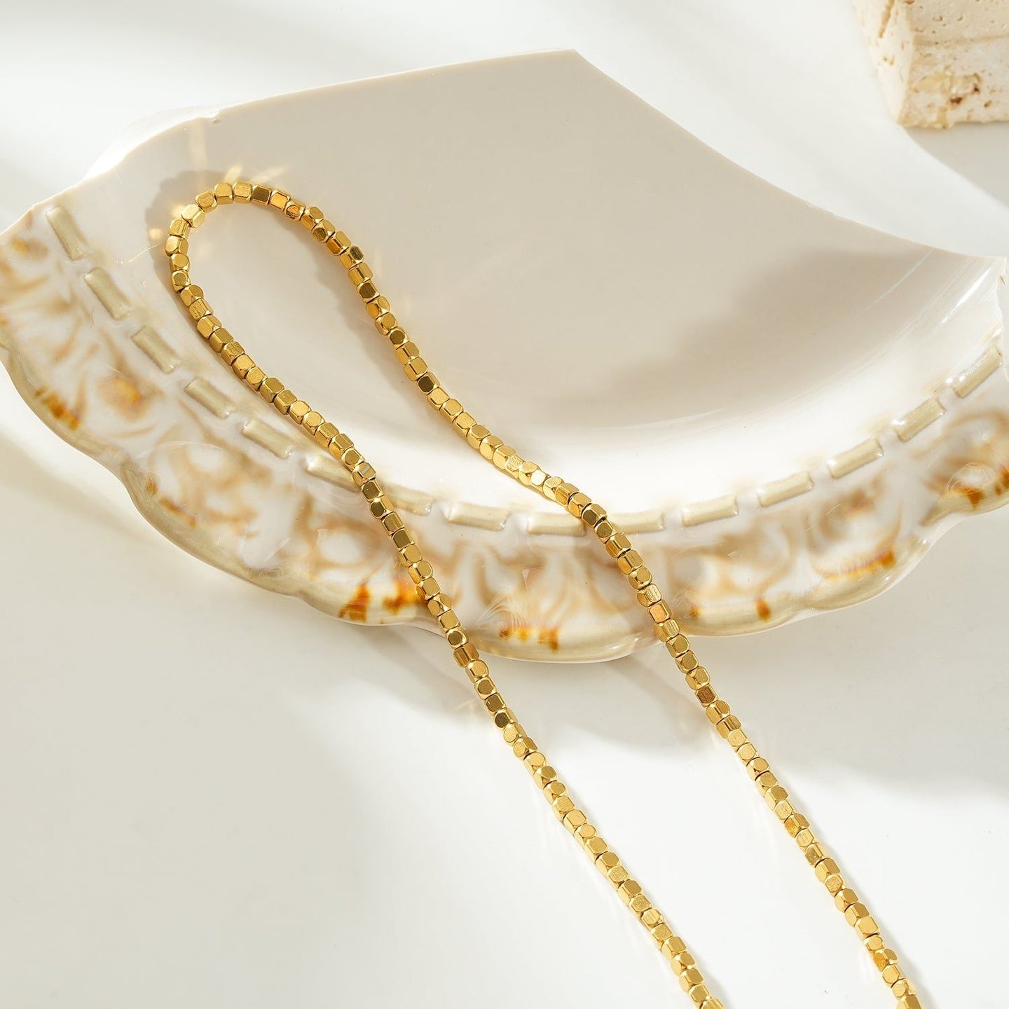 Gold beaded dainty chain necklace with small square beads, resting on a decorative ceramic dish with a scalloped edge, ideal for layering or everyday wear