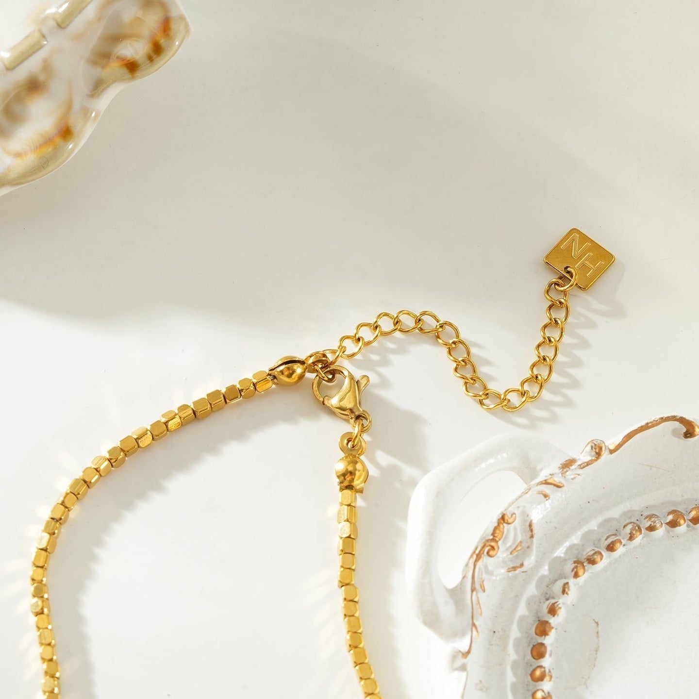 Gold beaded dainty chain necklace with small square beads, resting on a decorative ceramic dish with a scalloped edge, ideal for layering or everyday wear