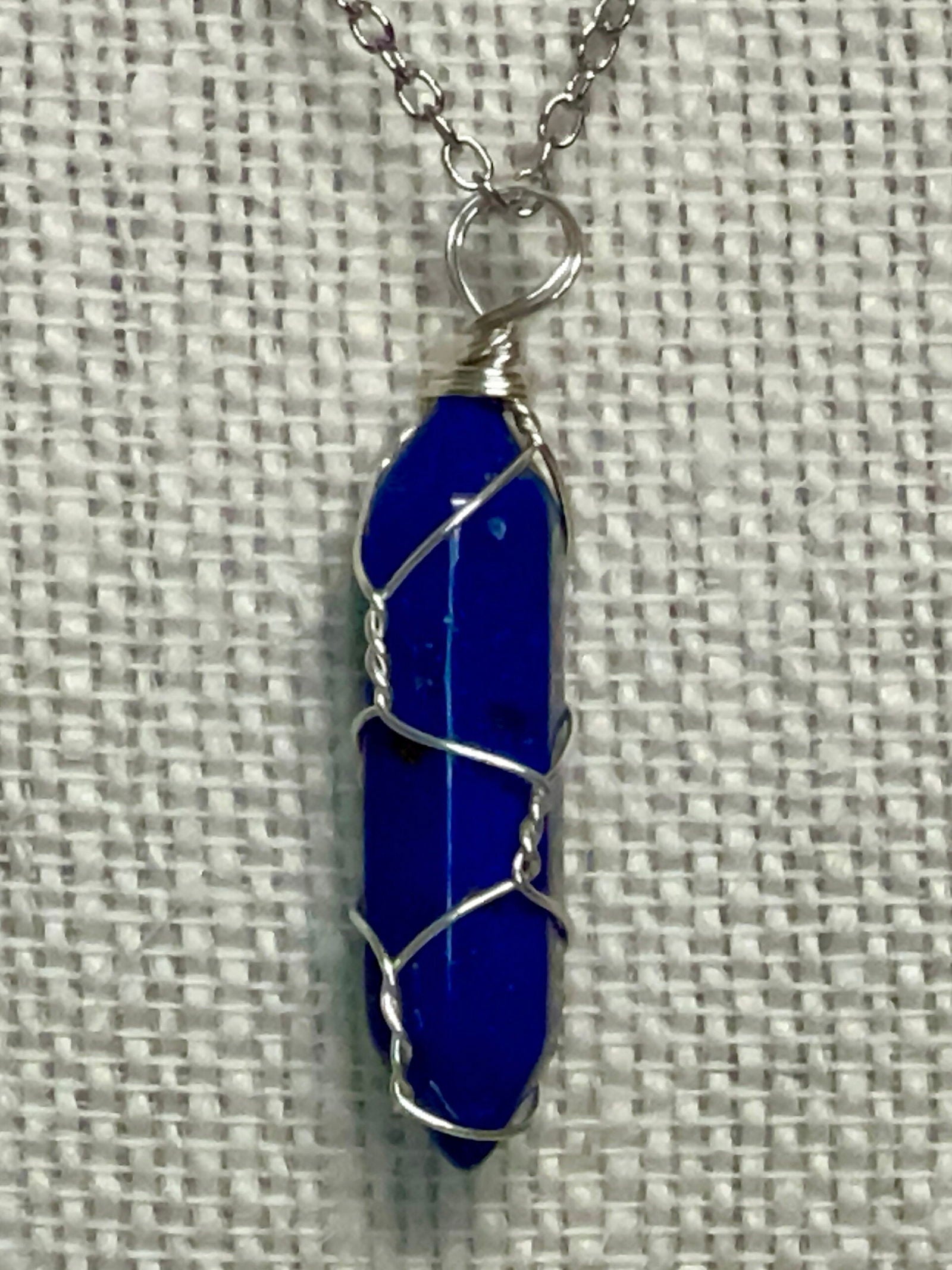 Silver chain necklace with a wire-wrapped deep blue gemstone pendant, set against a textured fabric background